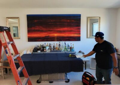A man stands by a covered table with bottles and tools in a modern room. A large sunset painting hangs on the wall behind the table. A ladder and packed boxes are nearby.