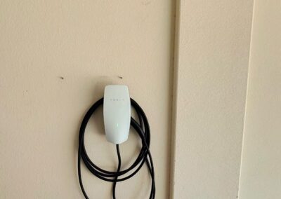 A Tesla wall connector with a coiled charging cable is mounted on a beige garage wall. Part of a metal rack is visible at the bottom left corner.