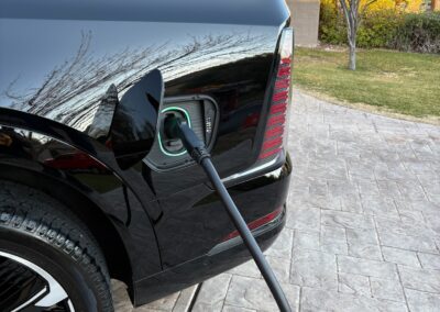 A black electric vehicle is parked on a stone driveway while plugged in and charging. Trees and grass are visible in the background on a sunny day.