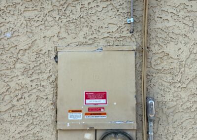 Beige electricity meter box with labels and a round glass meter mounted on a textured stucco wall, with metal conduit pipes running horizontally and vertically above and beside the box.
