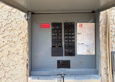 An open electrical breaker panel mounted on a textured beige wall, showing multiple circuit breakers, a faded inspection sticker, and a red warning label inside the metal door.