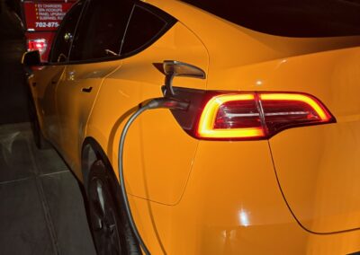 A yellow electric car is parked on a driveway at night, plugged into a charging cable. Its rear lights are illuminated, and a fire truck with flashing lights is visible in the background.