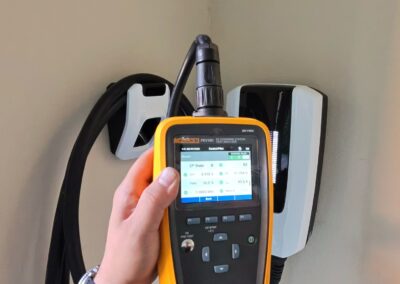 A hand holds a yellow and black electronic testing device in front of a wall-mounted electric vehicle charging station, with the chargers cable coiled on the wall behind.