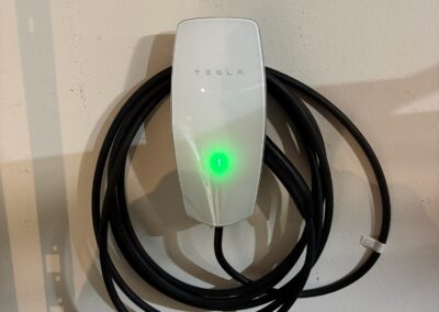 A white Tesla wall charger with a glowing green light is mounted on a wall. A black charging cable is coiled neatly around the charger.