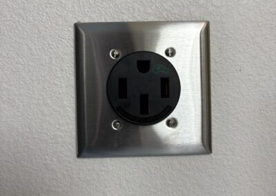 A close-up of a stainless steel electrical wall outlet with a round black socket, installed on a textured white wall. The outlet has three vertical prongs and is secured with four screws.