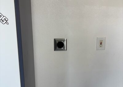 A wall with two fixtures: a black circular outlet with a metal plate and a standard white electrical outlet with two sockets and a red reset button. The left edge of a large object is also visible.