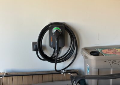 A black electric vehicle charging station with a coiled cable is mounted on a light-colored wall in a garage, next to storage bins and a power outlet.