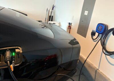 A black electric car is parked in a garage, plugged into a wall-mounted EV charger. A fire extinguisher, utility panel, and cleaning tools are visible on the wall in the background.