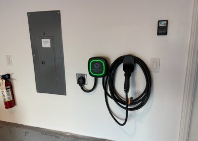 A home electric vehicle charging station is mounted on a white garage wall next to a gray electrical panel, a wall-mounted control device, and a fire extinguisher. The charging cable is neatly coiled.