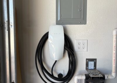 A Tesla wall connector is mounted on a garage wall, with its charging cable neatly coiled. Nearby are an electrical panel, double power outlet, and a blue water filter attached to white pipes.