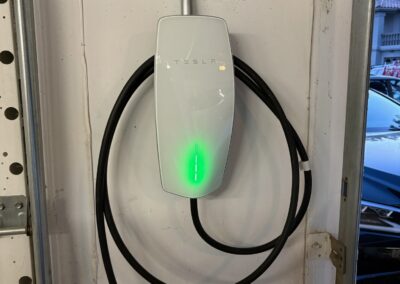 A Tesla wall charger with a glowing green indicator light is mounted on a garage wall, and its charging cable is neatly coiled around it. A blue vehicle is partially visible outside the garage.