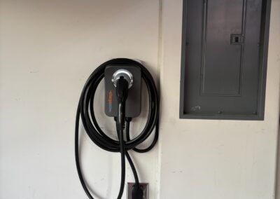 A wall-mounted electric vehicle charger with a coiled charging cable is installed next to a closed electrical panel on a white wall.