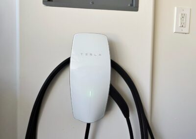 A Tesla wall charger with a coiled black charging cable is mounted on a white wall below an electrical panel and next to a power outlet. A small green light glows on the charger.