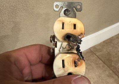 A hand holds a damaged electrical outlet with black burn marks and melted areas, indicating it has been scorched or experienced an electrical fire. The background shows a beige wall and tiled floor.