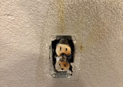 A damaged electrical outlet with exposed wiring is mounted on a textured wall. The wall around the outlet is stained and discolored, possibly from water damage or a previous leak.