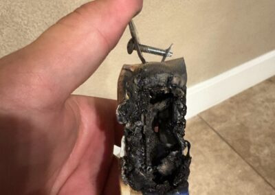 A hand holding a severely burned and melted electrical outlet with exposed screws; the outlet is blackened and charred, indicating significant fire damage.
