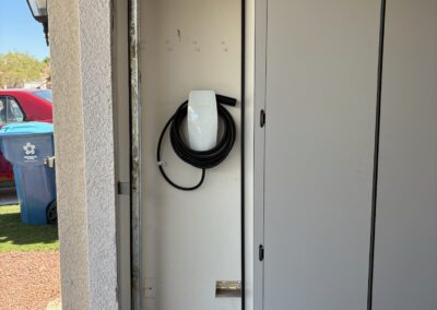A white electric vehicle charger is mounted on a garage wall, with its cable coiled. A tall white cabinet stands nearby. An open garage door reveals a blue car, a trash bin, and outdoor landscaping.