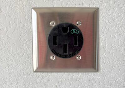 A close-up of a wall-mounted electrical outlet with three large prongs and a metallic cover, specifically designed for electric vehicle charging, as indicated by a green car symbol on the outlet.