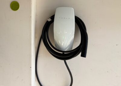 A white Tesla wall connector for electric vehicles is mounted on a white wall, with its black charging cable neatly coiled around it. A small green circle is visible on the upper left side of the wall.