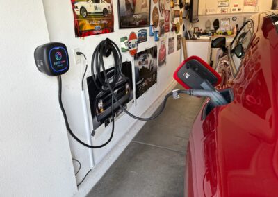 A red electric car is plugged into a wall-mounted charger in a garage decorated with various vintage car posters and memorabilia. The charger cable is connected to the car’s charging port.
