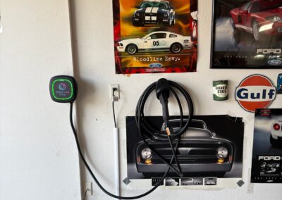 An electric vehicle charging station with a coiled cable is mounted on a garage wall, surrounded by posters of classic cars and automotive signs.