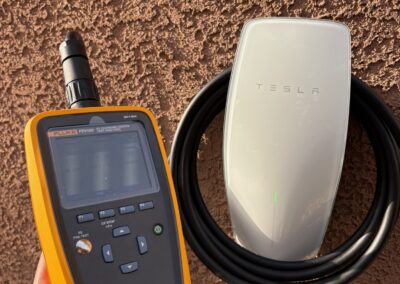 A hand holds a Fluke electrical tester next to a mounted Tesla wall charger with a coiled cable, both against a textured brown wall.
