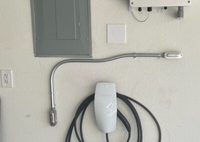 A wall-mounted electric vehicle charger with a coiled charging cable is installed below an electrical panel and near a white control box in a garage.