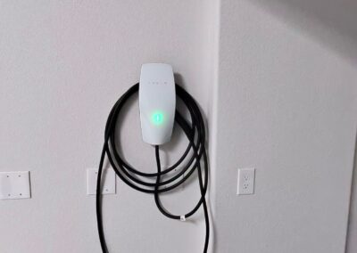A Tesla wall charger with a coiled charging cable is mounted on a white wall in a garage. The device displays a green light, indicating it is powered on. Several electrical outlets are visible nearby.