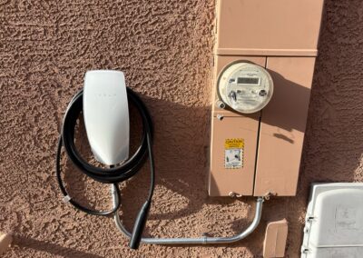 A Tesla electric vehicle wall charger is mounted on a textured exterior wall next to an electric meter and a caution sticker, with bright sunlight casting shadows.