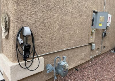 An electric vehicle charging station is mounted on a beige stucco wall next to a utility meter and several electrical panels, with gravel covering the ground below.