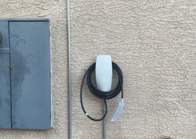 A Tesla electric vehicle wall charger with a coiled cable is mounted on a stucco wall next to a gray electrical panel and a utility meter. Some stone detailing is visible on the right side of the wall.