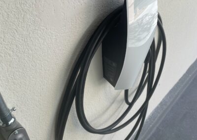 A wall-mounted electric vehicle charging station with a coiled black charging cable is attached to a light-colored wall near a dark floor.