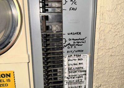 An electrical breaker panel with handwritten labels next to switches, including “A/C,” “FAU,” “Washer,” “G.F.I.,” and “Kitchen/Nook.” A warning sticker is partially visible on the left. The panel is installed on a textured wall.