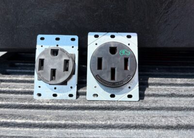 Two metal and black electrical outlets are mounted side by side on a textured surface. The outlet on the right has a green electric car symbol above the right slot. Both outlets have three slots.