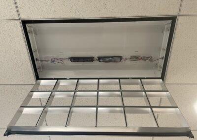 A ceiling light fixture with its cover open, exposing internal wiring and electrical components, is set in a white drop ceiling with square tiles. The light’s metal grid is hanging downward.