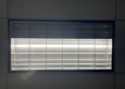 A close-up view of a rectangular fluorescent ceiling light fixture, illuminated and set into a tiled ceiling. The light is bright, with white grid panels diffusing the glow.