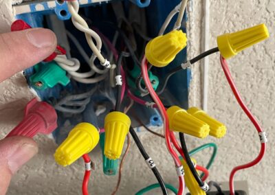 A close-up of an electrical wall box with multiple wires connected by red and yellow wire nuts. A persons hand is holding the wires, and the box is partially installed in a textured wall.