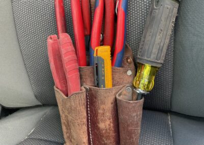 A brown leather tool pouch filled with various hand tools, including red-handled pliers, a yellow utility knife, and a screwdriver, resting on a gray fabric car seat.