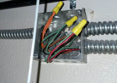 An electrical junction box with several colored wires (green, orange, black, and red) connected with yellow wire nuts, and metal conduit pipes attached to the sides, mounted on a beige wall.