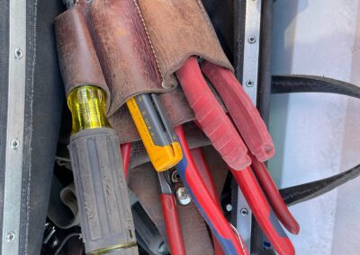 A leather tool pouch holds several red-handled pliers, a yellow-handled screwdriver, and a box cutter, all inside a metal-framed tool bag with other tools partially visible.