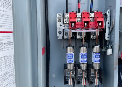 Electrical fuse box with three large fuses, red switches, black wiring, and green and yellow ground wires inside a metal enclosure. Instructions and labels are visible on the inside of the door.