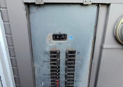 A metal electrical breaker panel is mounted on an exterior wall. The gray panel has two columns of black circuit breakers, some labeled with handwritten notes, and shows signs of wear and chipped paint.