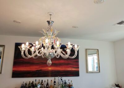 A modern dining room with a large glass chandelier, a table with packages, a sideboard with various bottles, two mirrors, and a sunset painting on the wall. Light streams in from a window on the right.