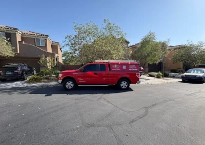 A bright red utility truck is parked on a residential street lined with modern, two-story houses and trees under a clear blue sky.